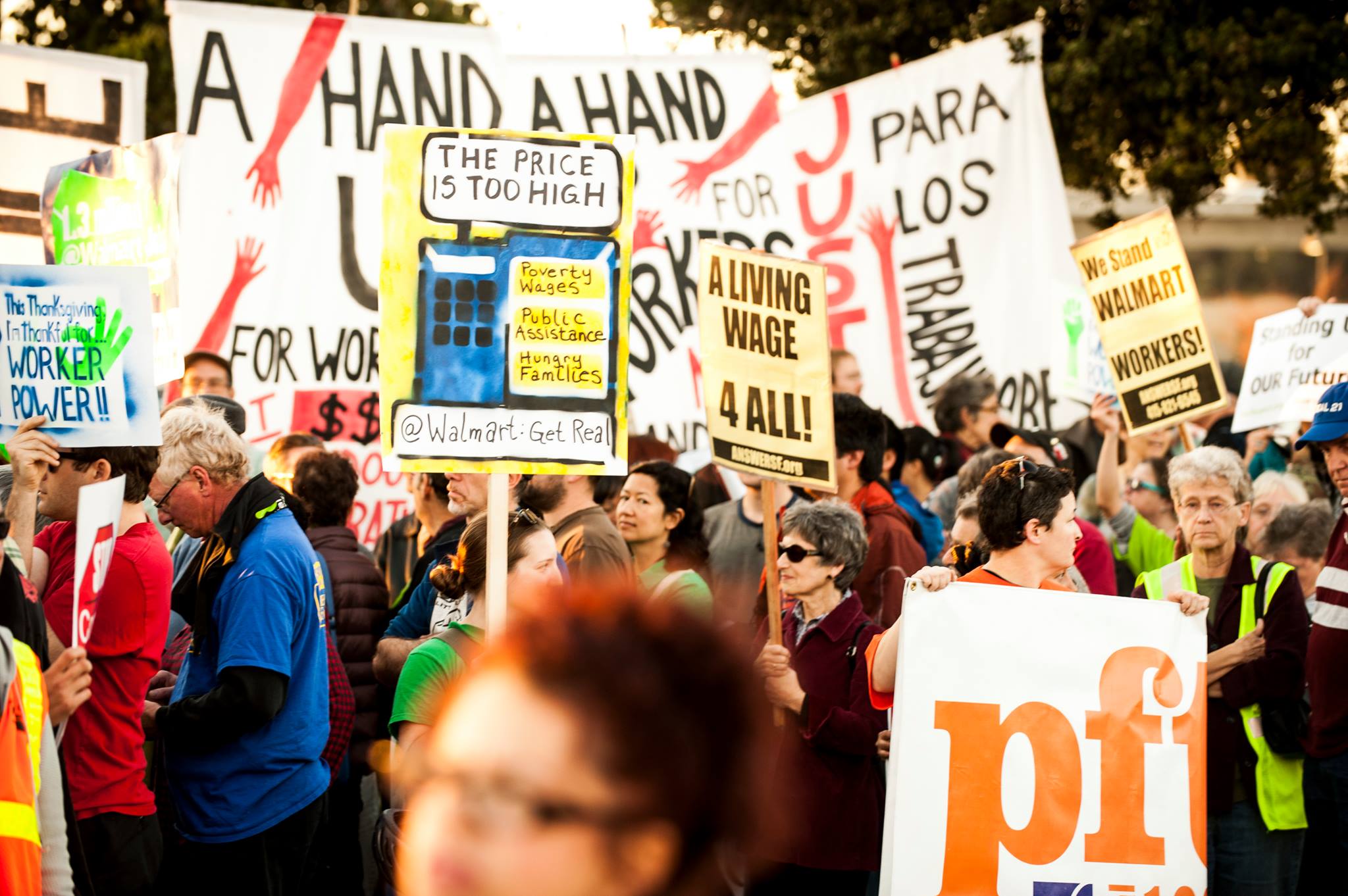Protesters in a crowd at a Walmart protest. Various signs read, "A living wage 4 all!", "The price is too high - poverty wages - public assistance - hungry families", and "This Thanksgiving I'm thankful for worker power!"