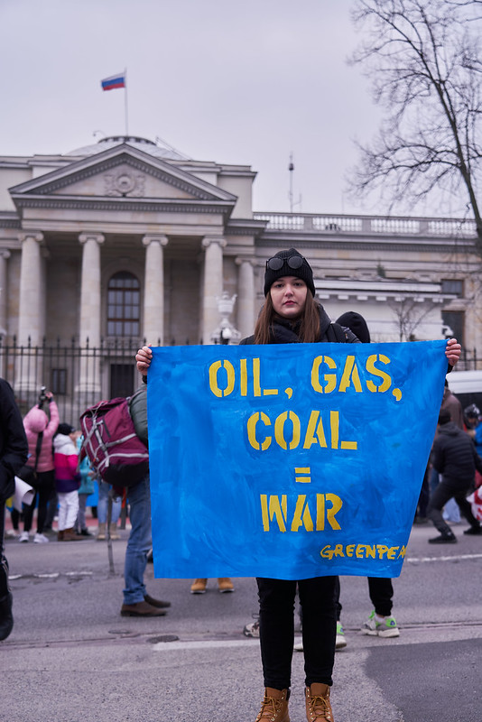 A photo of Paulina Gruda standing in front of a government building holding a sign that says "Oil, Gas, Coal = War" with the Greenpeace logo in the corner
